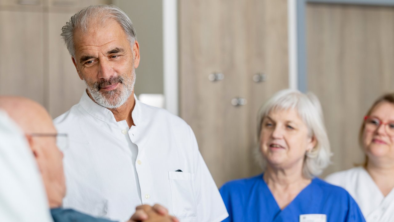 Ein Arzt spricht mit einem Patienten, der im Patientenbett liegt. Herum stehen 2 weitere Pflegerinnen
