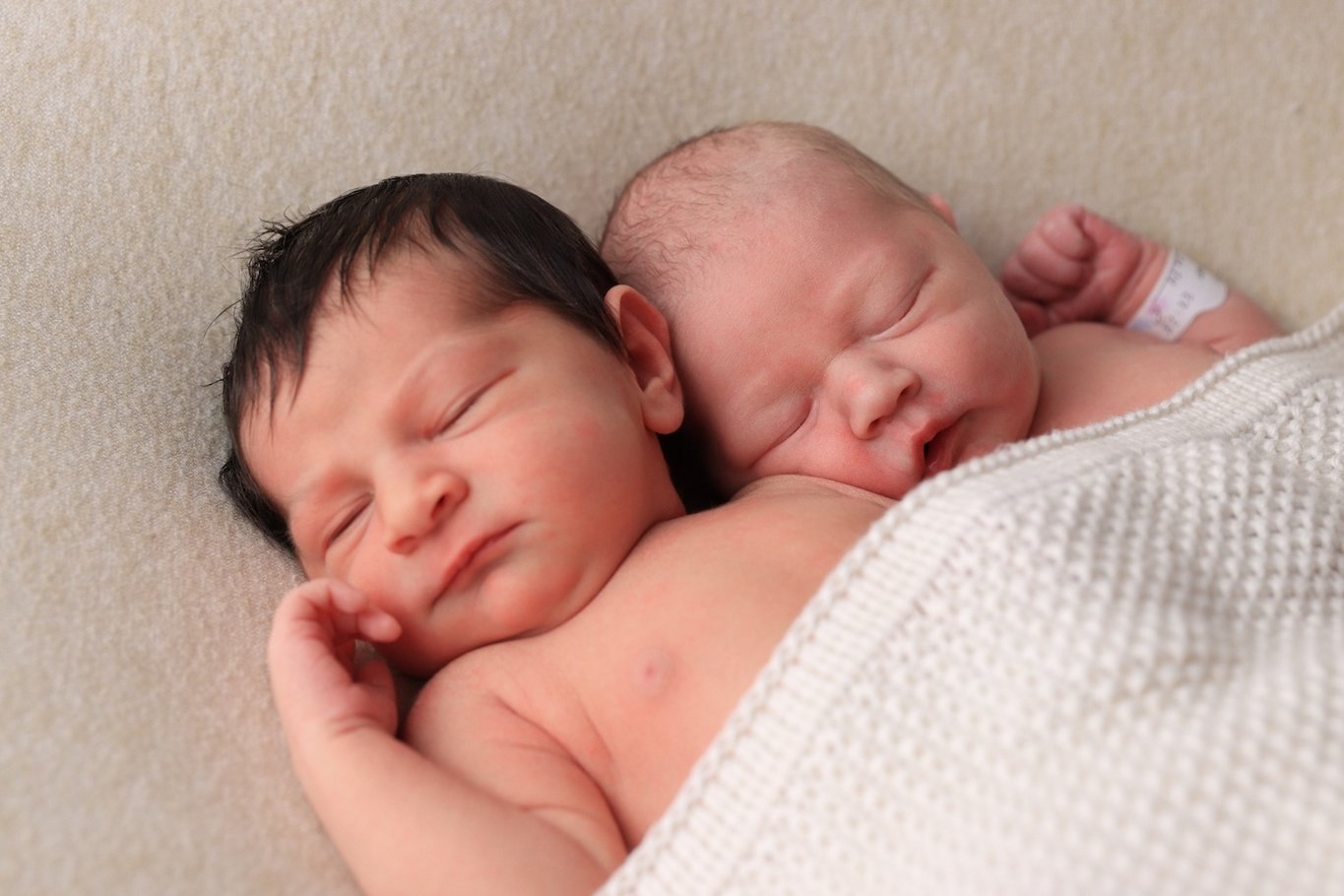 Zwei Neugeborene liegen eng aneinander gekuschelt und schlafen. Beide Babys sind in eine helle Decke gehüllt und liegen auf einer weichen Unterlage.