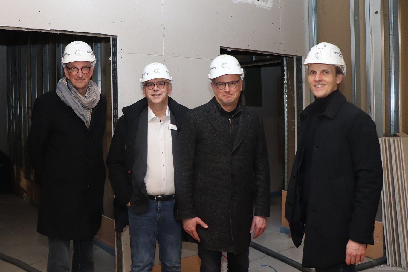 Vier Männer mit Schutzhelmen stehen auf einer Baustelle im Krankenhaus Reinbek in einem unfertigen Innenraum nebeneinander und blicken in die Kamera.
