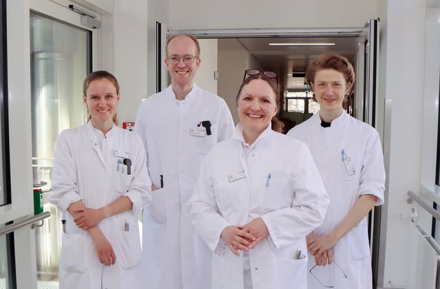 Vier Ärztinnen und Ärzte in weißen Kitteln stehen lächelnd im Flur eines Krankenhauses und blicken in die Kamera.