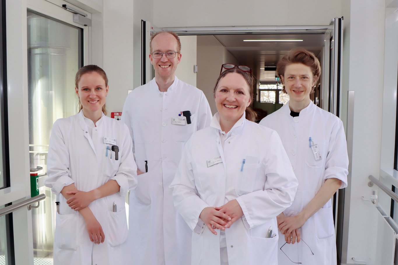 Vier Ärztinnen und Ärzte in weißen Kitteln stehen lächelnd im Flur eines Krankenhauses und blicken in die Kamera.