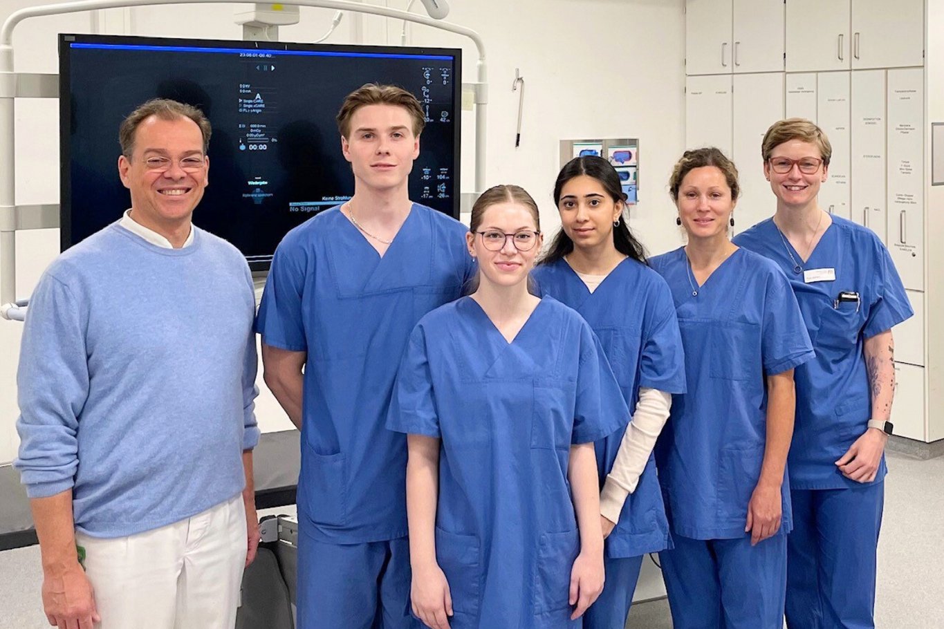Eine Gruppe von Mitarbeitenden und Auszubildenden in blauer medizinischer Arbeitskleidung steht gemeinsam in einem klinischen Raum. Die Personen blicken in die Kamera, im Hintergrund sind medizinische Geräte und Schränke zu sehen.