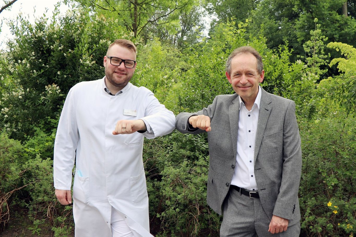 Zwei Männer stehen draußen vor Grünpflanzen und stoßen ihre Ellenbogen aneinander.