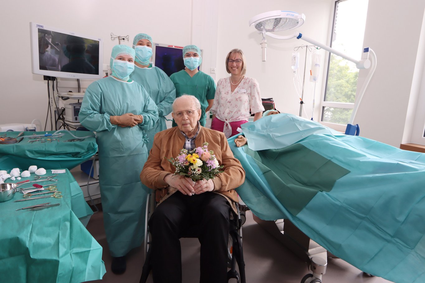 Ein alter Herr sitzt mit Blumenstraus in der Hand im Rollstuhl neben ihm zwei OP Azubis im OP-Simulations-Raum