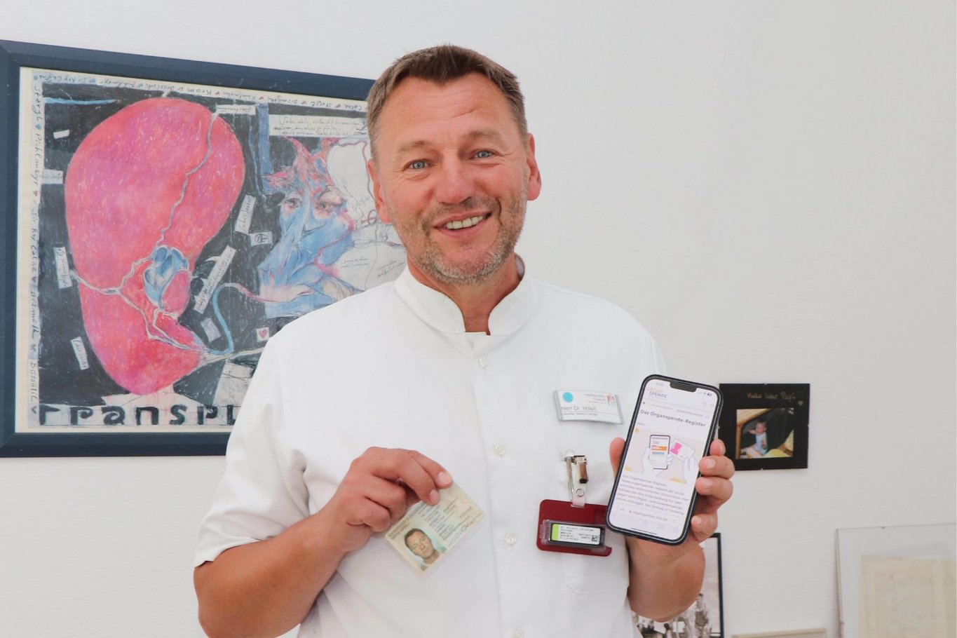 Arzt in weißer Dienstkleidung steht vor einer Wand mit medizinischer Illustration und hält einen Organspendeausweis sowie ein Smartphone mit Informationen zur Organspende in die Kamera.