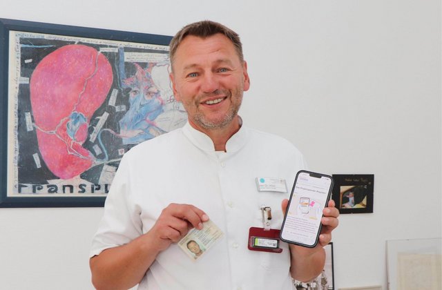 Arzt in weißer Dienstkleidung steht vor einer Wand mit medizinischer Illustration und hält einen Organspendeausweis sowie ein Smartphone mit Informationen zur Organspende in die Kamera.