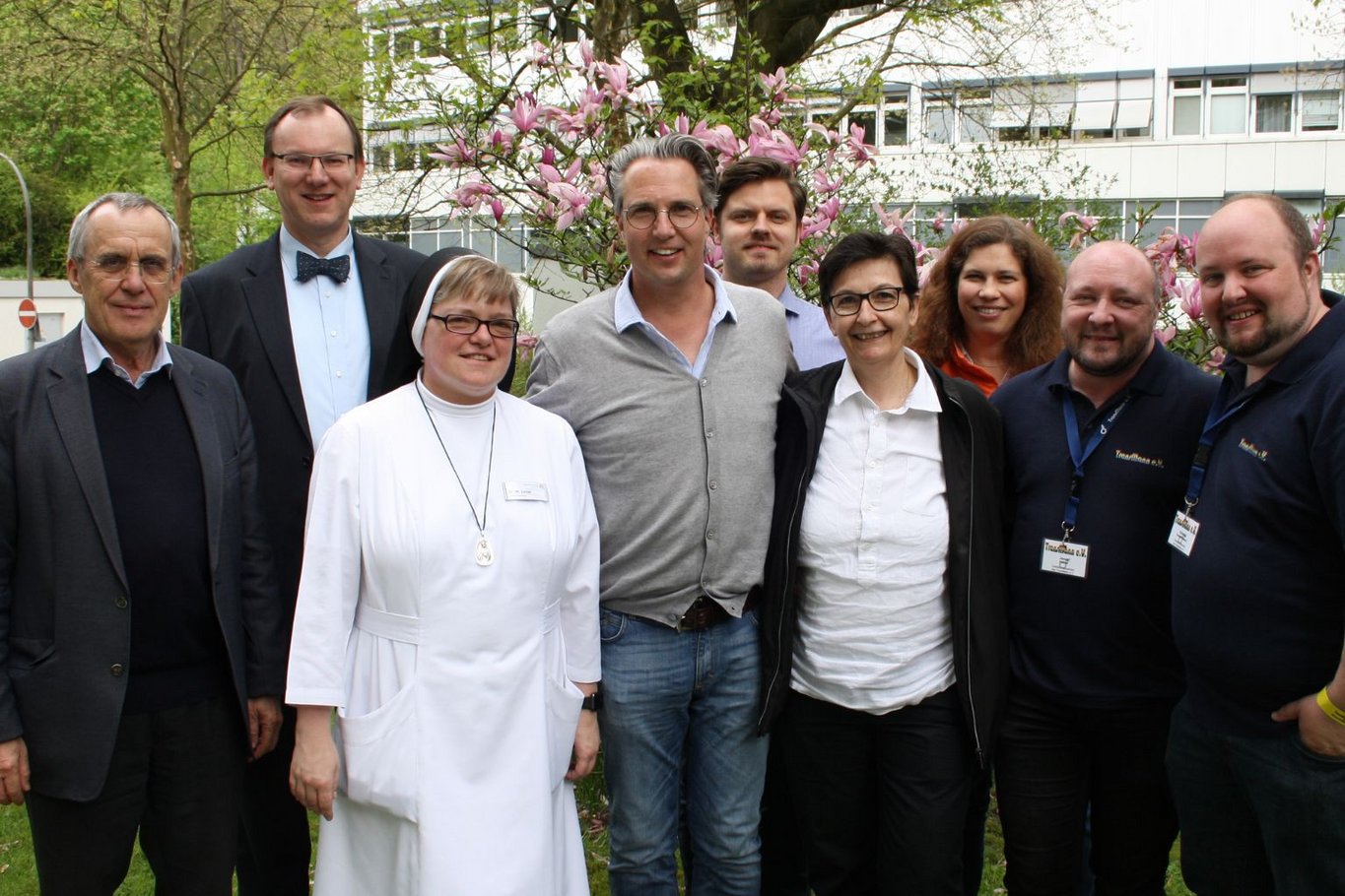Gruppe von neun Personen, darunter eine Ordensschwester und mehrere Männer und Frauen, stehen gemeinsam im Garten vor einem Gebäude mit blühendem Baum und lächeln in die Kamera.