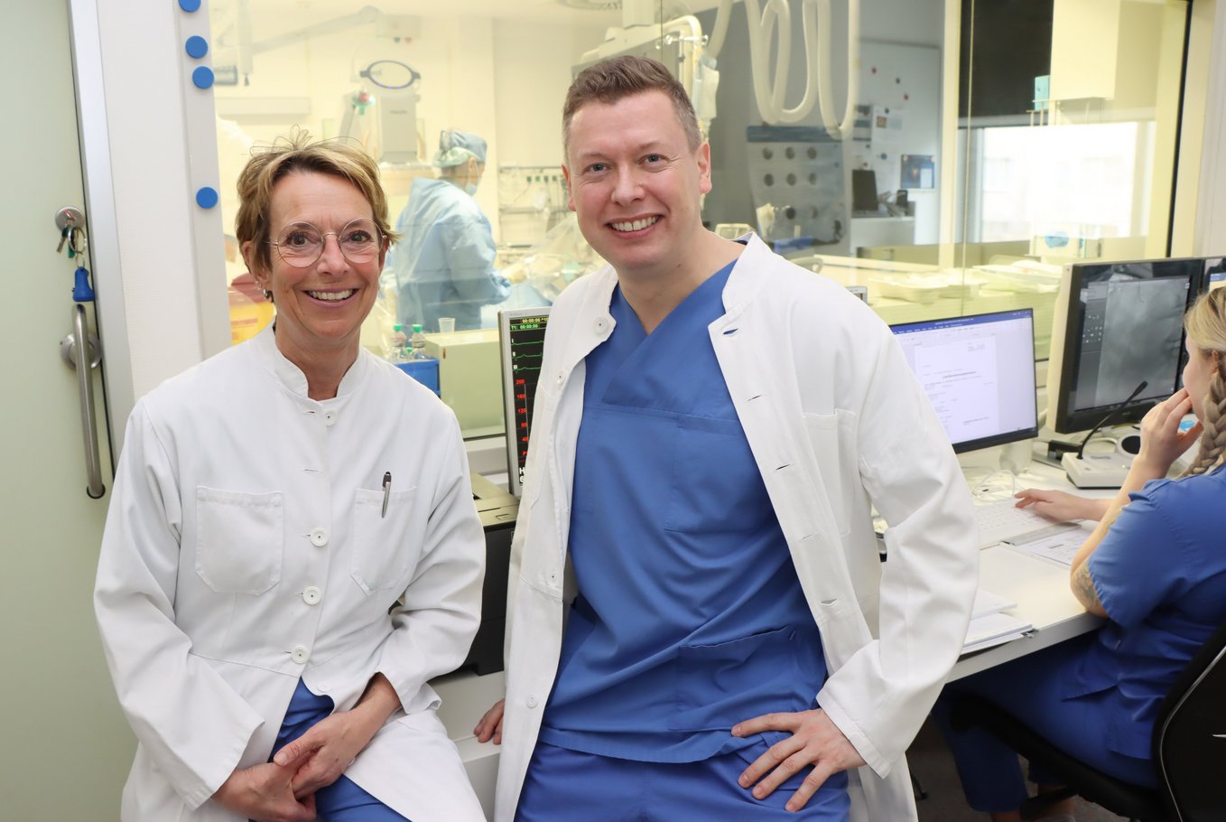 Eine Ärztin und ein Arzt stehen zusammen vor dem Herzkatheterlabor und lächeln in die Kamera