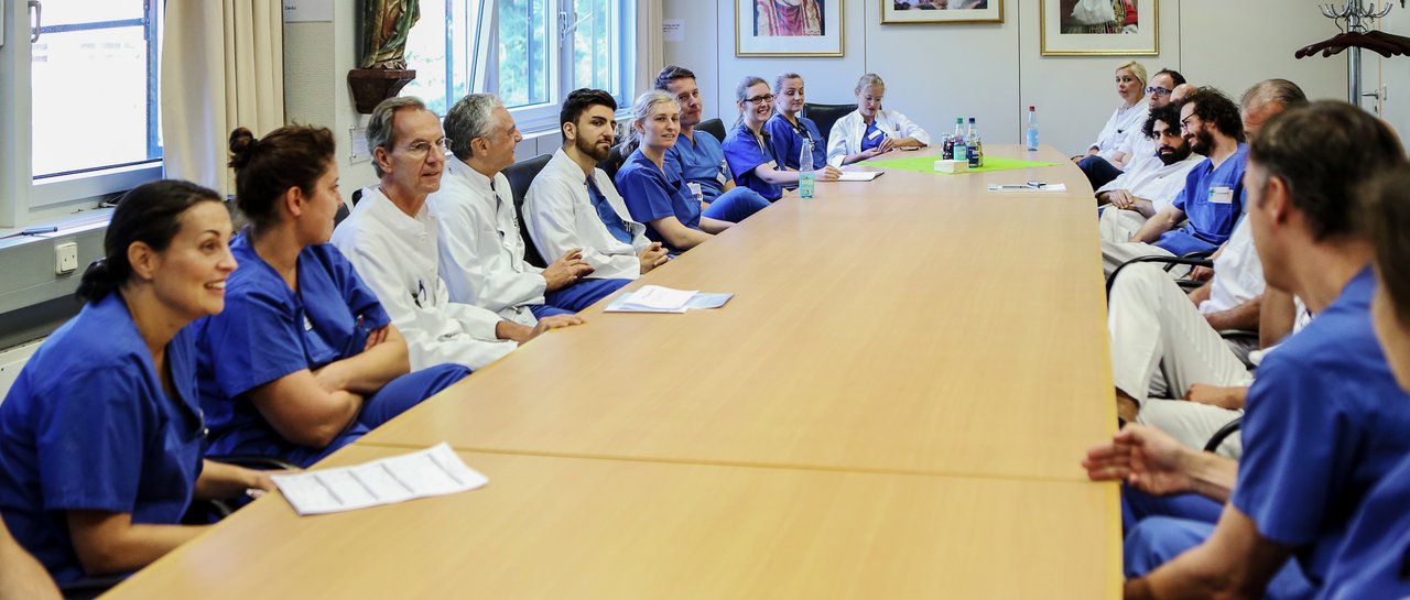 Teambesprechung von Ärzt:innen, alle sitzen an einem großen Tisch und diskutieren