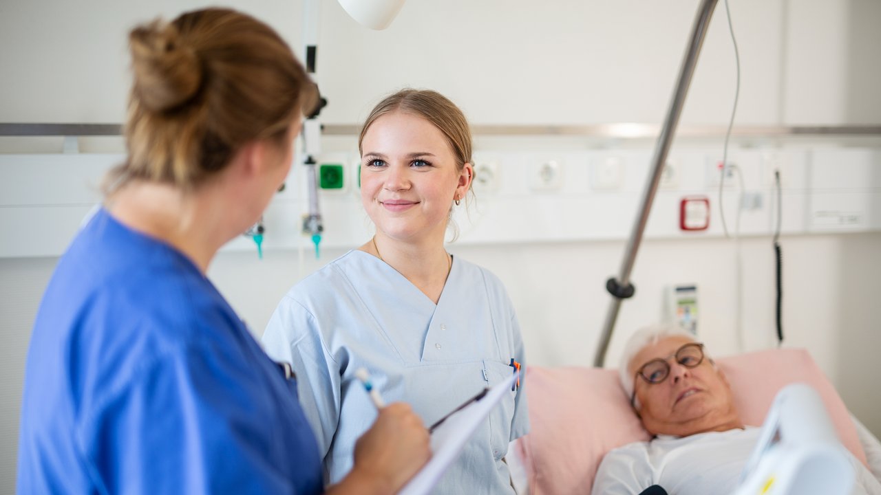Eine Praxisanleiterin spricht mit einer Azubine im Patientenzimmer, im Hintergrund ein Patient im Krankenhausbett
