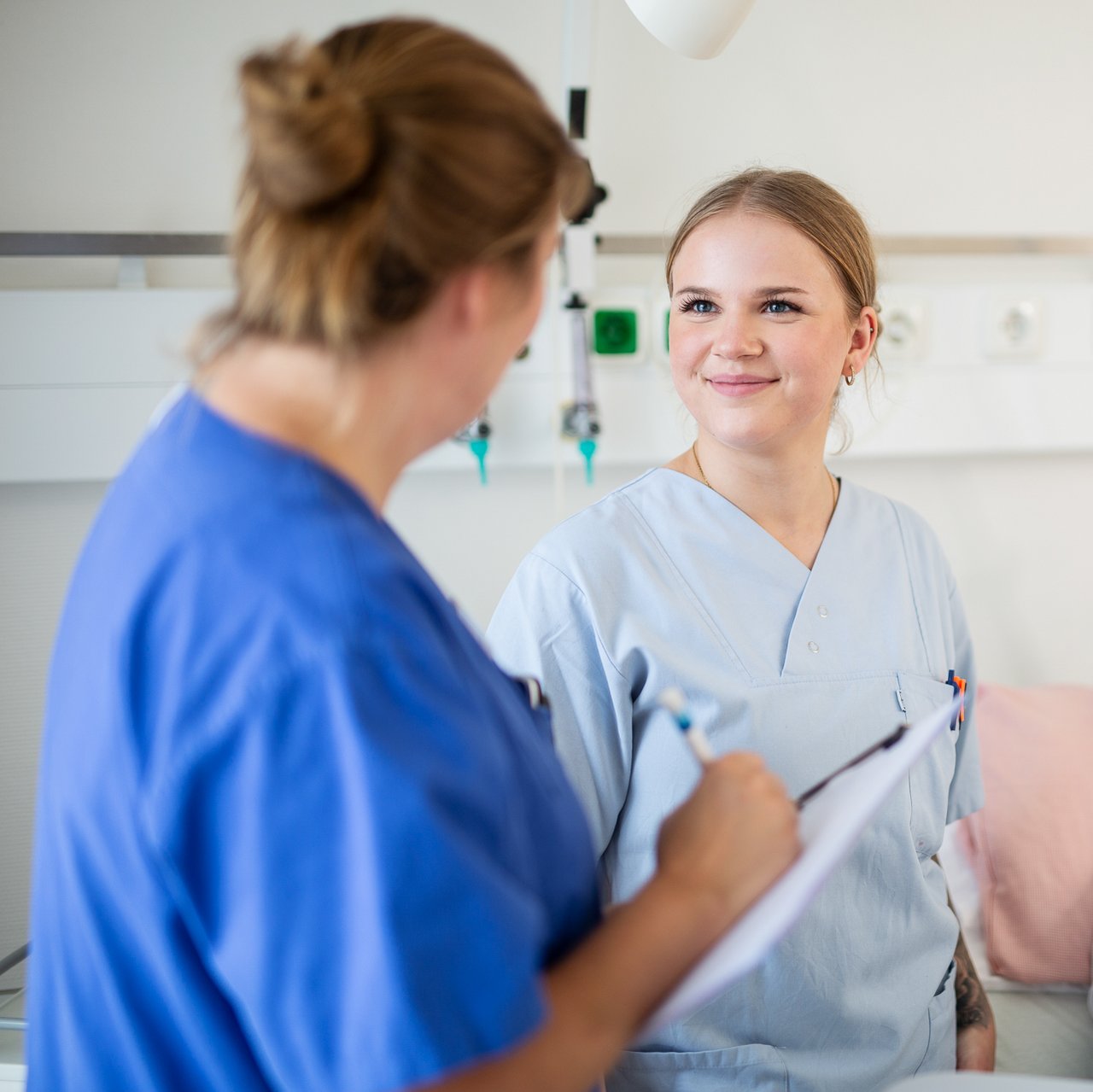 Eine Praxisanleiterin spricht mit einer Azubine im Patientenzimmer, im Hintergrund ein Patient im Krankenhausbett