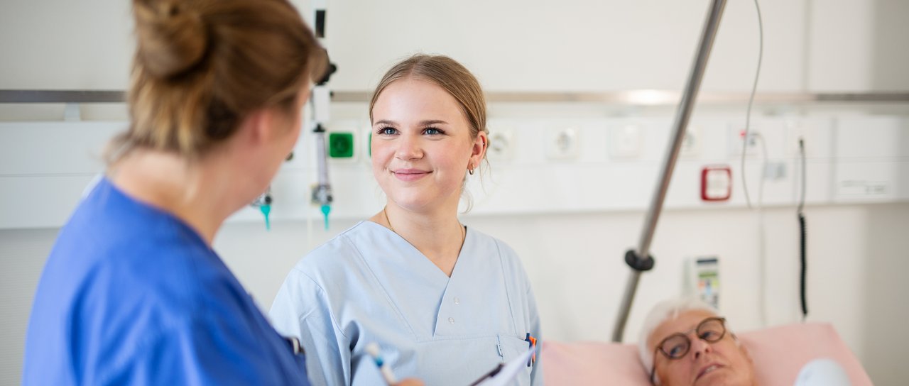 Eine Praxisanleiterin spricht mit einer Azubine im Patientenzimmer, im Hintergrund ein Patient im Krankenhausbett