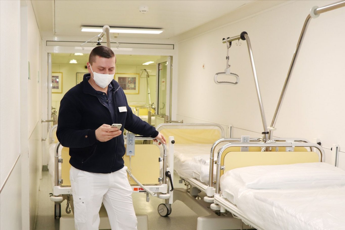 Ein Krankenhausmitarbeiter mit Mundschutz steht im Flur neben einem Patientenbett und schaut auf ein mobiles Gerät, über das er einen neuen Transportauftrag erhält.