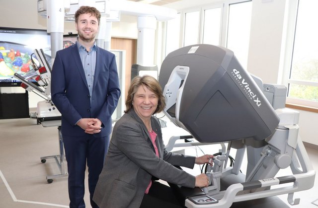 Gesundheitsministerin von der Decken sitzt an einem medizinischen Operationssimulator im Trainingsraum eines Krankenhauses, während ein Mann im Anzug daneben steht und beide in die Kamera lächeln.
