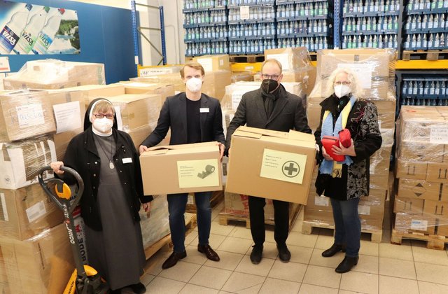Vier Personen stehen in einem Lager zwischen Paletten mit Hilfsgütern und halten Kartons mit medizinischem Material für ukrainische Flüchtlinge.