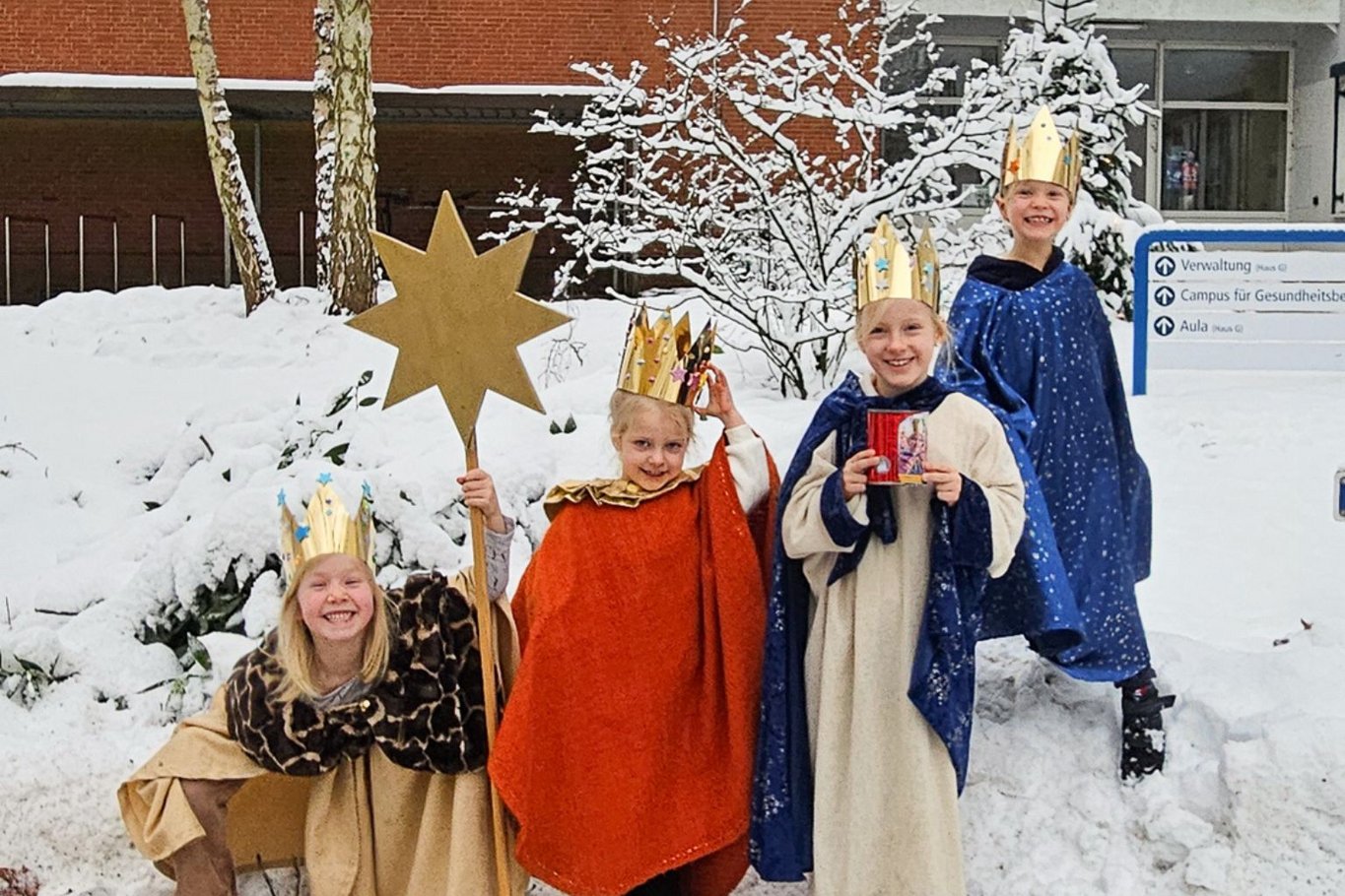 Vier als Sternsinger verkleidete Kinder stehen im Schnee vor dem Verwaltungsgebäude des Krankenhauses Reinbek; sie tragen Kronen und Gewänder, eines hält einen Sternstab und ein anderes eine Spendenbox.