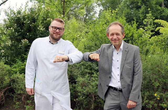 Zwei Männer stehen draußen vor Grünpflanzen und stoßen ihre Ellenbogen aneinander.