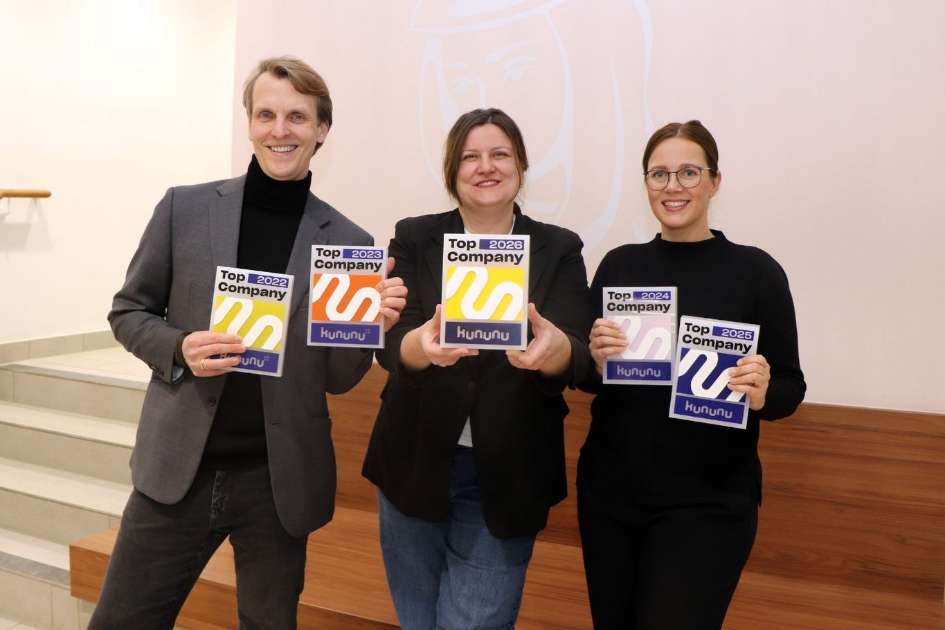 Drei Mitarbeitende des Krankenhauses Reinbek stehen nebeneinander und halten mehrere „Top Company“-Auszeichnungen der Arbeitgeberplattform kununu in die Kamera. Links steht Geschäftsführer Fabian Linke, in der Mitte Personalleiterin Ines Kerßenfischer und rechts Recruiterin Sarah Baudzus. Sie zeigen Auszeichnungen aus den Jahren 2022, 2023, 2024, 2025 und 2026. Die drei Personen stehen vor einer hellen Wand und lächeln in die Kamera.