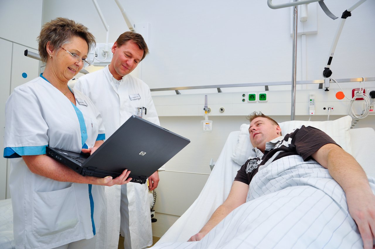 Eine Pflegerin und ein Arzt schauen auf einen Laptop neben ihnen ein Patient im Krankenhausbett