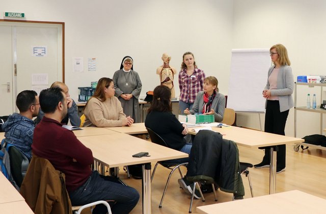Mehrere Kursteilnehmende sitzen in einem Klassenraum an Tischen und hören den Dozentinnen zu, die vorne stehen und den Unterricht eines berufsbezogenen Sprachkurses leiten; im Hintergrund sind ein Flipchart und ein Anatomiemodell zu sehen.