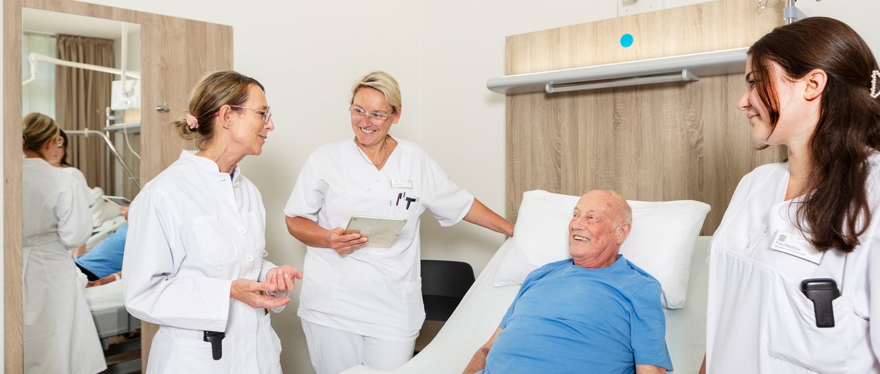 Die Chefärztin und ein 2 Pflegerinnen der Klinik für Kardiologie sprechen mit einem Patienten, ein älterer Herr, der im Patientenbett im Krankenhaus liegt.
