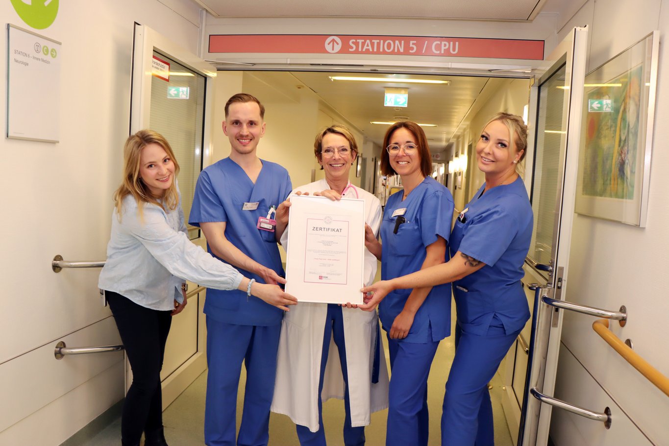 5 Personen, ein Mix aus Pflege Ärzte und Verwaltungsmitarbeiterin, halten stolz ein großes Zertifikat hoch im Krankenhausflur