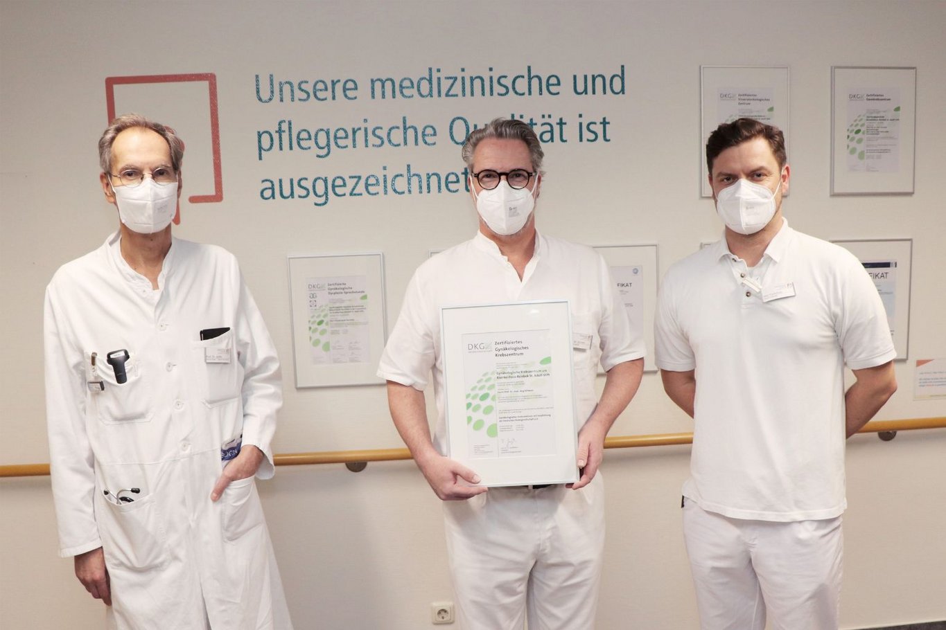 Drei Ärzte mit Mundschutz stehen vor einer Wand mit Zertifikaten, während einer von ihnen ein Zertifikat für das gynäkologische Krebszentrum in den Händen hält.