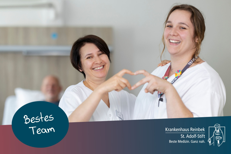 Zwei Pflegerinnen formen zusammen ein Herz mit den Händen und lächeln in die Kamera