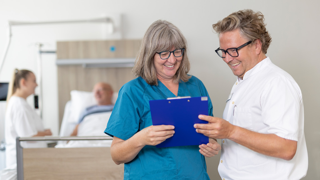Dr. Dick und Bertschart stehen vor einem Patientenbett und besprechen etwas, das sie auf dem Tablet sehen