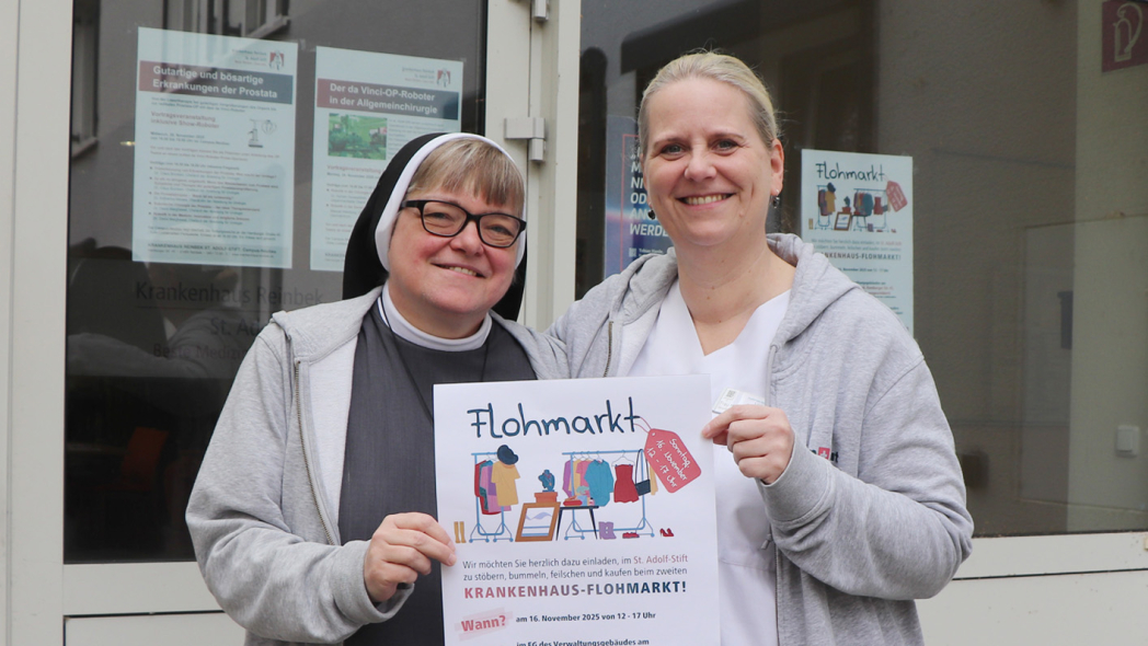 (v.l.n.r.) Schwester Luise und Yvonne Hillebrand vom Organisationsteam zeigen vor dem Eingang der Aula das Plakat mit der Werbung für den Flohmarkt am 16. November im St. Adolf-Stift.