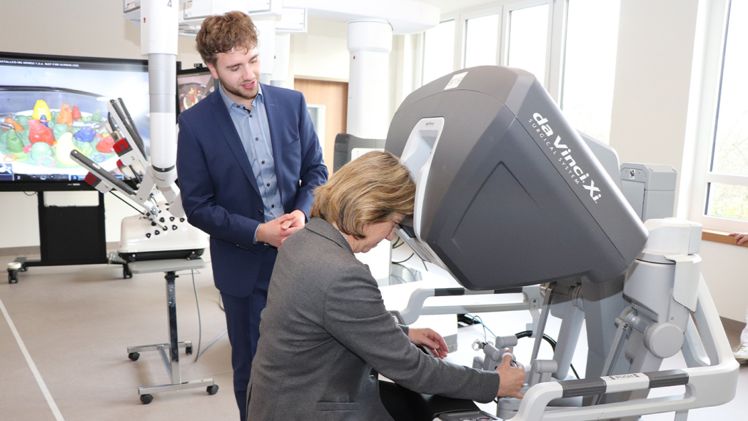 Bei einem Rundgang durch den Neubau erklärte OTA-Auszubildende Maximilian Weise der Gesundheitsministerin Kerstin von der Decken einen Operationsroboter, der eigens für die Veranstaltung im Campus aufgestellt wurde. Er lud sie ein sich an die Steuerkonsole zu setzen, um selbst zu erfahren, wie intuitiv die Arme zu bedienen sind. Ein baugleicher OP-Roboter steht nur 100 Meter weiter im Zentral-OP des St. Adolf